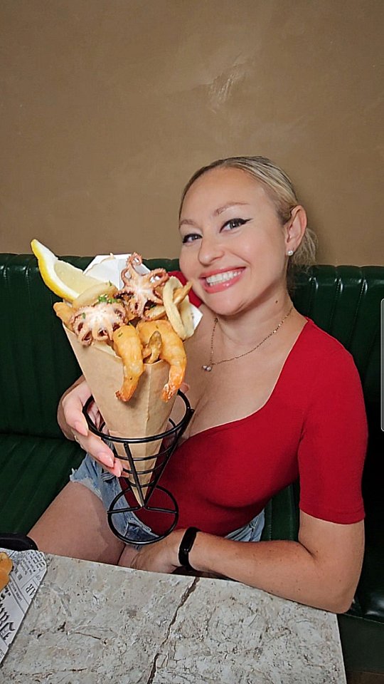 A woman with blonde hair and a red top is holding a paper cone filled with fried seafood, including shrimp, octopus, and fries. She is smiling, and the background shows a green booth at a table with a marble surface.