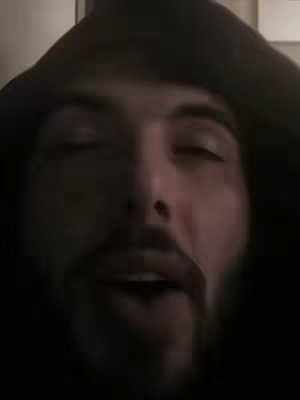 A close-up shot of a man wearing a dark hoodie, gazing towards the camera with his eyes closed, mouth slightly open. The lighting appears dim, casting shadows and highlighting his facial features, including his beard.