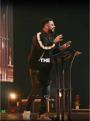 A man in a black jacket with white stripes with the word "THE" in white text in the image. The man appears to be giving a speech behind a podium with a microphone in hand.