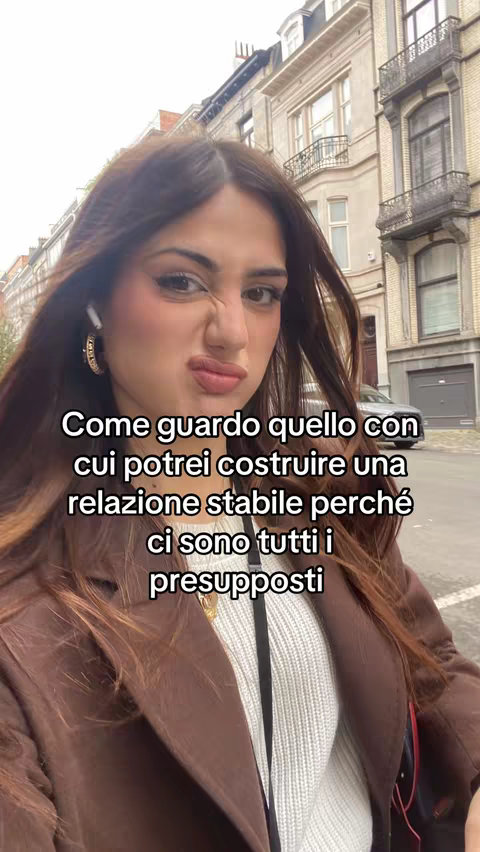 A selfie-style image featuring a woman in an outdoor setting with light-colored buildings in the background. The woman has dark hair and is wearing a brown coat over a white sweater. She has a slightly puckered expression, and text overlays the image.