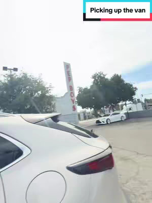 A POV shot from inside a car, showing the rear of a white vehicle in the foreground and a blurred view of the surroundings. The sky is bright. A sign that says RED BOYS is visible. The text overlay 'Picking up the van' is set within a white and black rectangular bar.