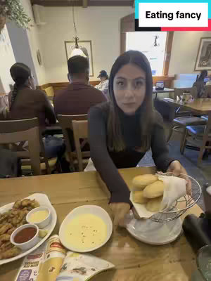 A woman sits at a restaurant table in a well-lit environment. She is holding a basket of rolls and looking at the camera. Various plates of food and sauces are on the table, along with a magazine and restaurant setting visible in the background.