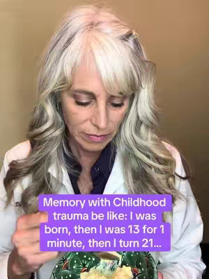 A woman with long, wavy gray hair and makeup is looking down with a contemplative expression. There is a purple rectangle containing text overlaid on the image. She is wearing a white coat over a dark blue top. She appears to be holding a decorative bowl in her hands. The background is a neutral, possibly indoor, setting