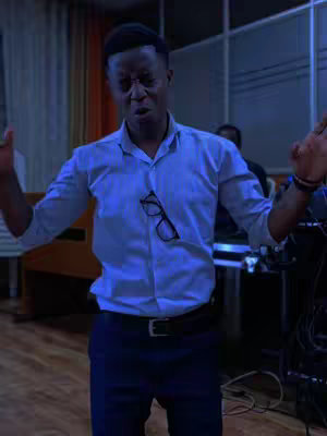 A man stands with arms raised in a room, possibly a stage or studio, wearing a light blue button-down shirt and dark pants. He has glasses clipped to his shirt.