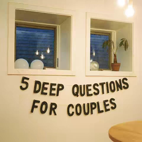 The image features a well-lit interior with two square windows flanking a wall where a banner reading '5 DEEP QUESTIONS FOR COUPLES' hangs. The windows have white frames and recessed shelves. The windows display a view of a blue sky with light, and decor on the shelf beneath the windows.