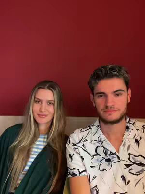 A medium shot shows two people, a woman on the left and a man on the right, against a dark red background. The woman is wearing a green top with a striped shirt underneath. Her blonde hair is parted in the middle. The man is wearing a white shirt with a black floral pattern. Both are looking at the camera.