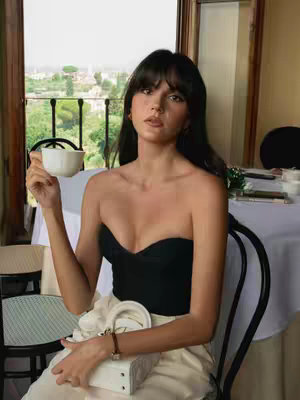 A young woman with long dark hair and bangs, wearing a black strapless top and cream-colored skirt, sits in a chair with a white cup in her left hand, and her right hand is in her lap holding a white purse. She is looking at the camera. She is sitting in front of a window that overlooks a European-style landscape.