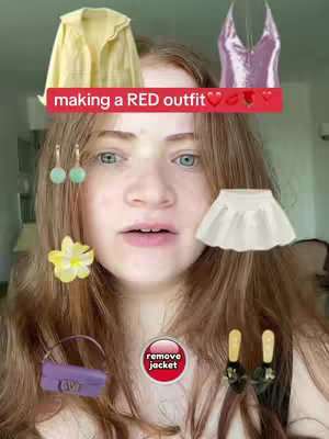 A selfie-style image featuring a young woman with long auburn hair and fair skin. She has a slightly open mouth and is looking towards the camera. Floating on top of her head are a yellow jacket and a pink camisole. There's a text overlay across the top that reads 'making a RED outfit'. Several other clothing accessories, including earrings, a skirt, a handbag, and shoes are scattered around. The top of the scene has a green wall in the background.