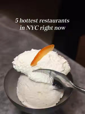 The image shows a close-up of a bowl of ice cream with a scoop being taken out of it. The ice cream is white, and a slice of orange is placed on the top. The bowl appears to be metallic and a spoon is used to take an ice cream scoop out. In the background, a dark, blurred setting is visible.