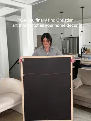 A woman stands in a bright, modern home, holding a large framed piece of art with its back facing the viewer. The frame is light wood, and the back is black. The woman is centered in the frame, with a neutral-toned living room visible in the background, including a white sofa and a glimpse of a kitchen with white cabinets.