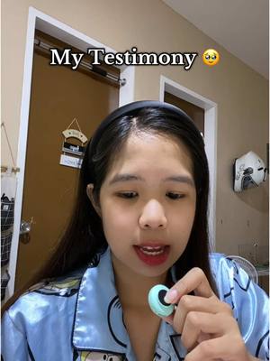 A woman with long dark hair wearing a black headband, pajamas, and red lipstick is holding a small, round, teal object. She's looking at the object while speaking. Above her, overlaid on the image, is the text 'My Testimony' with a surprised emoji. The background is a room.