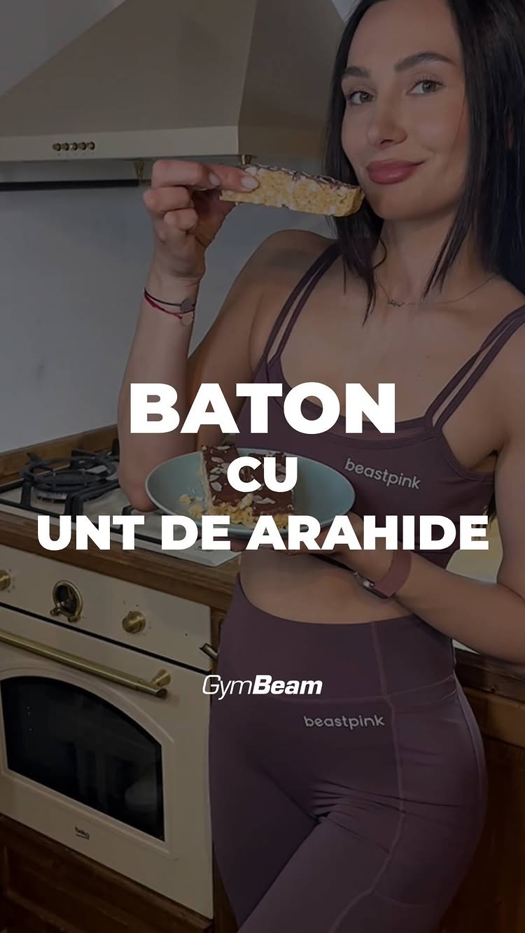A woman in workout attire is holding a piece of peanut butter and nut bar, smiling at the camera in a kitchen setting, with recipe title text over the image.