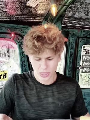 A young man with light skin and curly blond hair is captured in a selfie-style shot, with his face positioned prominently in the middle of the image. His eyes are slightly closed and his mouth is open. He appears to be speaking. He wears a dark, textured t-shirt. The background is a blurred depiction of an interior space, with green tones and what appears to be writing on the walls.