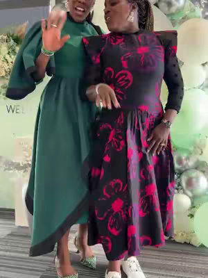 Two women are standing and smiling, seemingly at an event. One woman is wearing a dark dress with large pink floral patterns and white shoes. The other woman wears a green dress with a matching headwrap. The background shows white and silver balloons, possibly suggesting a celebration or party.
