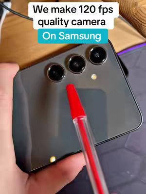 The cover image shows a close-up of a gray Samsung smartphone held with the camera lenses pointed towards the viewer. The top portion includes a rectangular white text overlay with a blue underline that reads "We make 120 fps quality camera On Samsung". A red pen is strategically placed to point towards the camera lenses. The background has a desk with a charging cable.