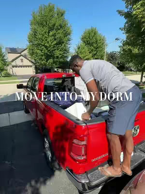 A man in casual attire unloading a bright red pickup truck parked outdoors. The clear blue sky and green trees in the background suggest a sunny day. The man is standing on the truck's bumper, reaching into the truck bed, which contains a white container and various items.