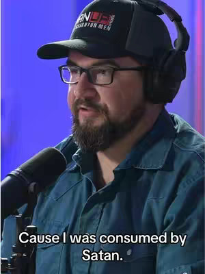 A man wearing a cap and glasses is in the center, speaking into a microphone. Text at the bottom reads, 'Cause I was consumed by Satan.' The background is a blurred blue light.