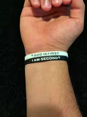 A close-up shot of a wrist wearing two silicone bracelets. The top bracelet is white with black text reading "GOD IS FIRST" and a small grey cross on each end of the writing. The bottom bracelet is black with white text that says "I AM SECOND" with a small grey cross on each end of the writing. The wrist appears to belong to a person with light skin. The background is a dark, blurry surface.