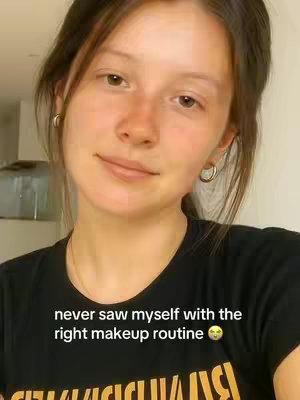 A selfie-style shot featuring a young woman with fair skin and brown hair styled away from her face. She's wearing a black t-shirt featuring a design partially visible at the bottom. The background is softly out of focus, likely a room setting, and a silver canister is seen on the upper left. The woman is looking at the camera with a slight smile and hoop earrings.
