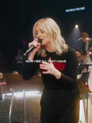 A woman with blonde hair is singing into a microphone on a stage. She is wearing a black shirt with a red graphic and black pants. The background is dark, with band members and stage elements. The lighting is focused on the woman.