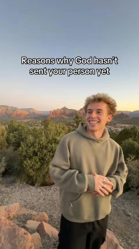 A young man with curly hair and a casual green hoodie smiles at the camera against a scenic backdrop of mountains and greenery under a sunset sky, with text overlayed on the image.