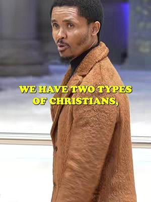 A man with short black hair and a beard is looking slightly to the side. He is wearing a brown coat over a dark turtleneck. The background is blurred.