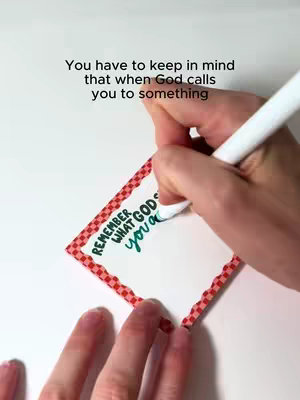 A close-up shot shows a hand writing on a white card decorated with a red and white patterned border. Above the card, there's text that reads, "You have to keep in mind that when God calls you to something." On the card, there are handwritten words in a teal color that say "REMEMBER WHAT GODS YOU a". The background is a clean, white surface.