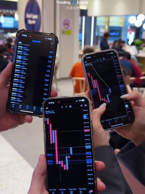 An image shows three hands holding smartphones. Each phone displays a chart with red and blue lines on a black background. The phones are held in front of a blurred background of what appears to be a public space with multiple people.