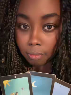 A close-up shot featuring a young woman with dark skin and braided hair. She is looking directly at the camera with a neutral expression. Her eyes are dark and expressive. She holds three tarot cards with illustrations of a moon, a star, and a sun in front of her. The background is a dark, possibly indoor setting.