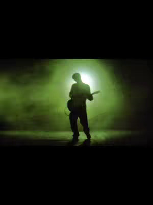 A silhouette of a person playing an electric guitar is illuminated by a bright green light, with a misty green background. The figure is fully blacked out against the light with a microphone cable hanging slightly.