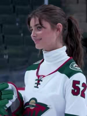 A young woman in a hockey jersey smiles to the side, the background is a hockey rink, she has a ponytail. She appears to be blowing a kiss.