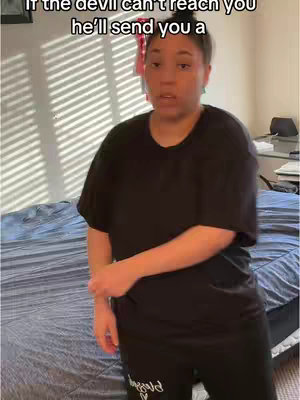 A woman in a black t-shirt and pants stands facing the camera in a bedroom. A bed with a blue cover is visible behind her along with a partially visible window and blinds.
