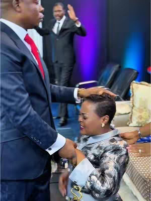 Image shows a formal setting with three people primarily in focus. A man in a dark suit and red tie is seen from the chest up, holding the head and hands of a woman in a patterned top and silver accessories. Behind them, a man in a dark suit has his arms raised in a gesture, with other individuals and chairs visible in the background.