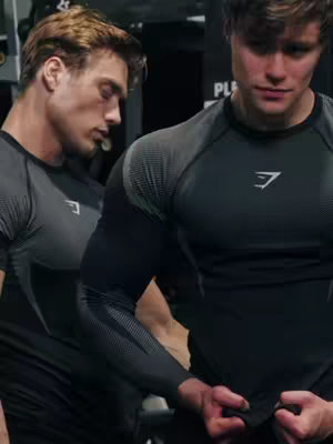 Cover image features two men in athletic wear, posing in gym. The one on the left is seen from the side, arms at his side, head slightly down, while the one on the right is facing the viewer, bending his arm, with a Gymshark logo visible. The color palette is dark, the background appears to be a gym setting.