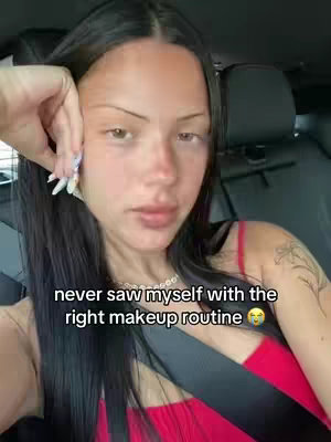 A woman is sitting in a car, looking at the camera. She has long black hair and is wearing a red top. The background is blurred, but it appears she is in the driver's seat. There is text overlayed on the image.