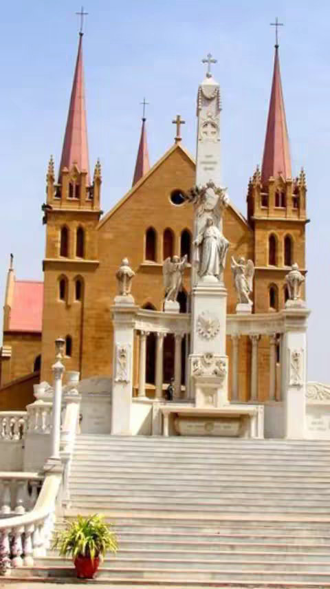 The image showcases a grand church building. The architecture is elaborate, with a facade of light-colored stone. The building features multiple spires with crosses atop them. There are steps leading up to the entrance, and statues are positioned on the facade.