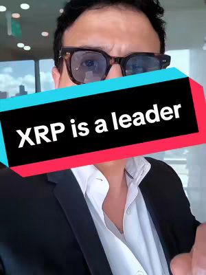 A man wearing glasses and a white shirt is speaking in front of a camera. A banner with the text 'XRP is a leader' is placed in front of him. The background is blurred, suggesting an indoor setting.