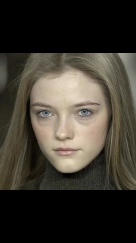 Close-up shot of a young woman with fair skin, light blonde hair, and blue eyes. She is wearing a dark turtleneck. The background is blurred, suggesting a soft, professional aesthetic.