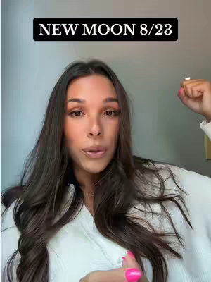 A woman is centered in a medium shot, gazing directly at the camera. Above her head, a black rectangular banner displays the text 'NEW MOON 8/23'. She has long, dark brown hair, styled with a slight wave, framing her face. Her complexion has medium skin tone with brown eyes and full lips, with a visible makeup look. She wears a white robe and pink nail polish on what is visible of her right hand. The background is a gradient grayish-white, slightly blurred. She seems to be speaking or about to speak.