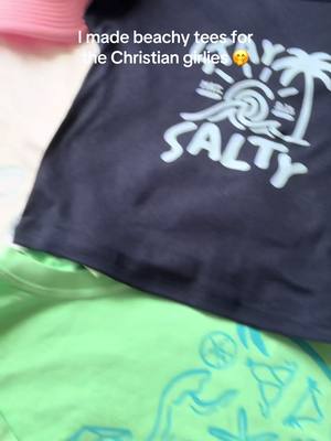 The image showcases three t-shirts of different colors: pink, navy blue and green, arranged in a vertical stack. The navy blue shirt in the middle features a white graphic with the word 'Salty' prominently displayed underneath a design of a sun and palm tree. The green shirt at the bottom and the pink shirt at the top are partially visible, with the pink shirt appearing to have no visible design and the green shirt suggesting an abstract pattern. The text overlay reads, 'I made beachy tees for the Christian girlies'.