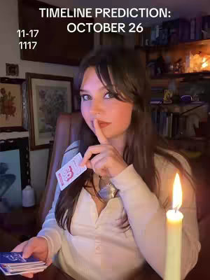 A woman with long brown hair is seated indoors, holding a tarot card and gesturing for silence. The background includes framed pictures and a lit candle, with text overlays indicating a prediction.