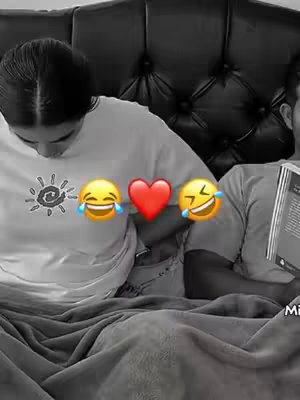 Black and white image of a couple lying in bed. The woman is looking down, and there are emojis on her t-shirt. The man is reading a book. A bedspread covers them.