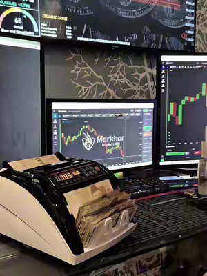 The image shows a desk filled with monitors displaying financial charts. A money-counting machine is in the foreground with a stack of bills. The overall composition suggests a focus on trading or financial activities.