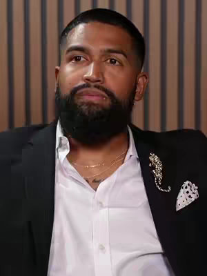 A medium shot of a man of color in a suit, seated and looking slightly upwards and to the left. He has a neatly trimmed black beard and short, styled hair. He is wearing a white shirt with the top button undone and a black suit jacket. A gold chain necklace sits just above the unbuttoned shirt. The jacket includes a decorative brooch and a pocket square.