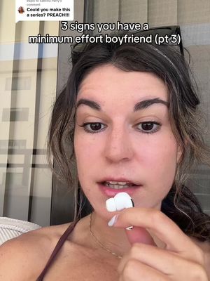 A selfie-style shot featuring a woman holding an AirPod in her hand, with text overlayed on the image. The text reads "3 signs you have a minimum effort boyfriend (pt 3)". The woman is facing the camera directly, with a slight upward angle. The background is blurred, but appears to be a room interior.
