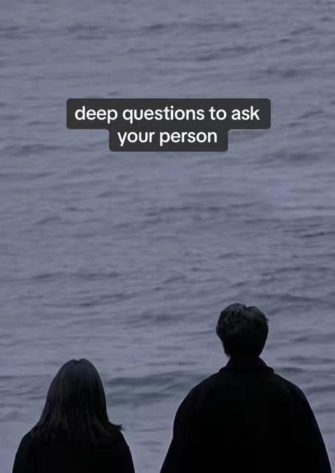 Two people seen from the back standing on a shore, looking out at a body of water. The sky is overcast and the water a greyish blue. A text overlay states, "deep questions to ask your person".
