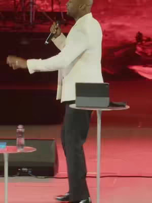 A man in a white suit jacket stands on a stage holding a microphone, with red lighting in the background. There's a table with a laptop on it in the foreground.