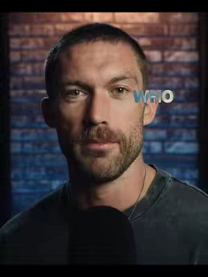 A man with short brown hair, a beard, and a serious expression is framed from the chest up. Over his right shoulder the word “WHO” appears in a turquoise color. The background is a dark blue brick wall.