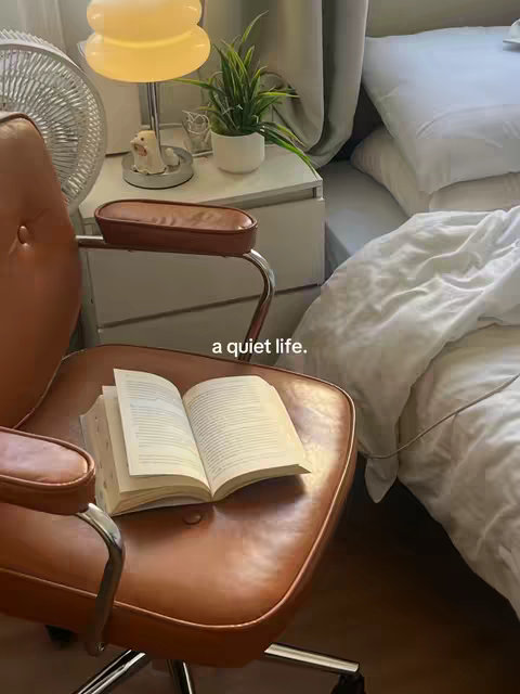 A cozy interior scene featuring a room dominated by neutral tones and soft light. The focal point is a tan leather armchair with an open book on its seat, suggesting reading. A bed with white bedding occupies the background on the right side. Additional decorative elements, such as a lamp, a small plant, and a fan, add to the relaxed atmosphere, reinforcing themes of tranquility and solitude. The overall lighting is soft, creating a calm and inviting ambiance.