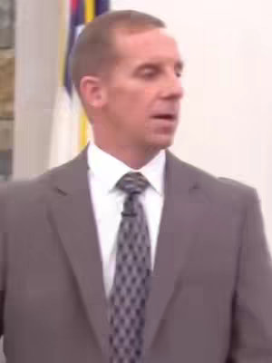 A medium shot showing a man in a suit standing in front of a blurred white background. He is facing slightly to the left and appears to be speaking. He is wearing a gray suit, a white shirt, and a patterned tie. The lighting seems professional.